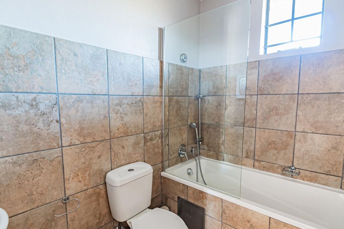 A functional bathroom featuring a bathtub with a glass shower screen, a toilet, and neutral-toned ceramic wall tiling.