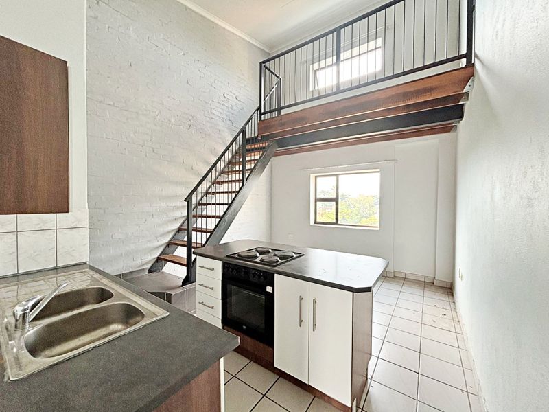 Open-plan kitchen and living area featuring a mezzanine level, exposed brick wall, built-in oven, electric hob, and tiled flooring throughout.