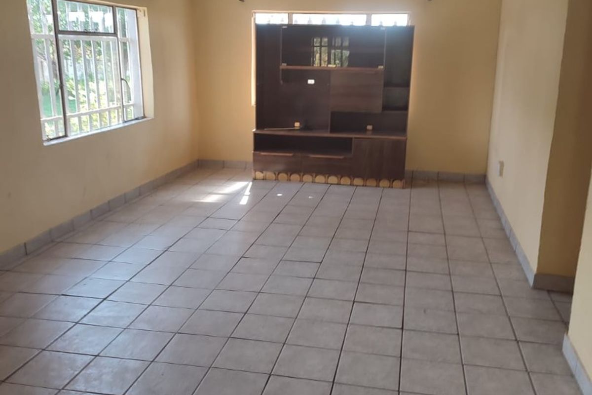 A spacious, empty living room featuring neutral ceramic floor tiles, security bars on windows, and a glossy ceiling finish.