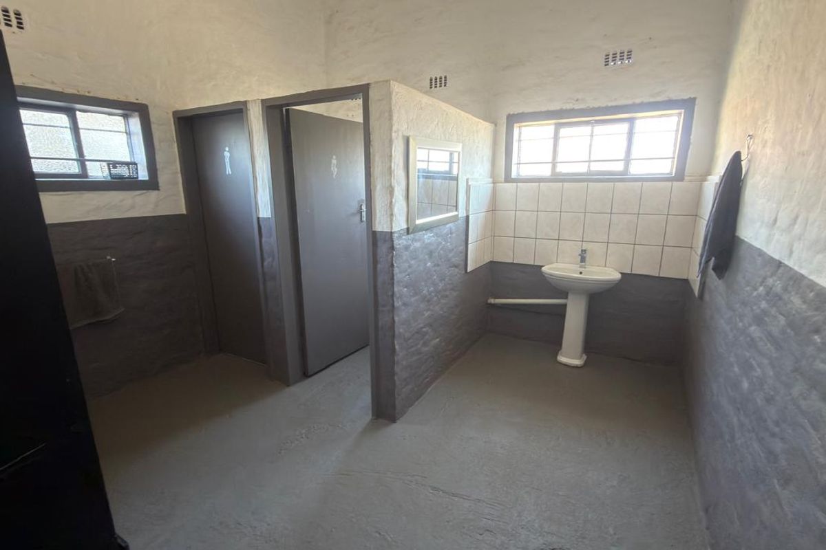 A functional commercial restroom facility featuring a pedestal basin, tiled splashback, and two separate toilet cubicles with painted brick walls.
