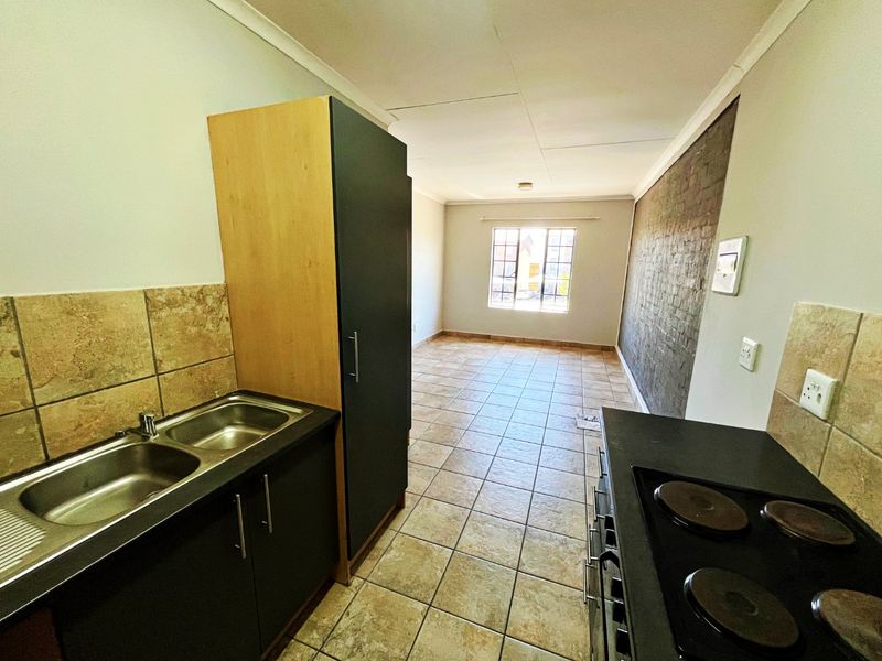 An open-plan kitchen and living area featuring tiled flooring, a double sink, built-in cabinetry, an electric hob, and an exposed brick feature wall.