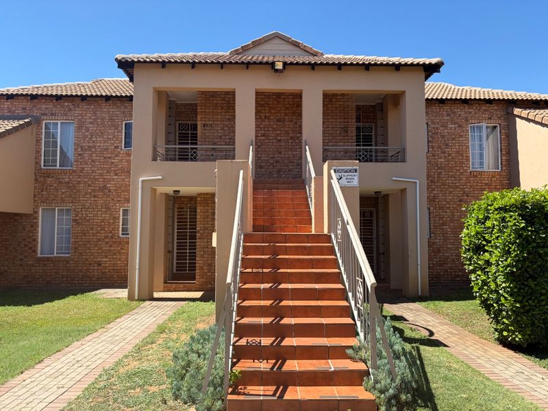 Front elevation of a two-story brick apartment complex featuring an external staircase, tiled roof, and paved walkways leading to the building entrances.