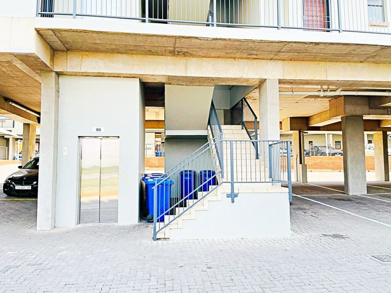 Ground level apartment building entrance featuring a modern elevator, concrete staircase with metal railings, and covered parking area.