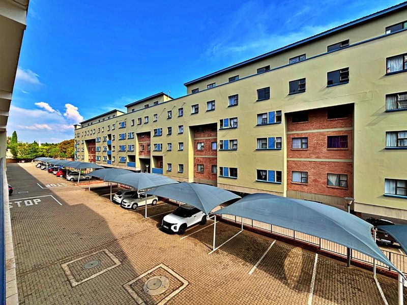 Exterior view of a multi-story apartment complex featuring a paved parking area with shade cloth carports and a well-maintained brick and plaster facade.