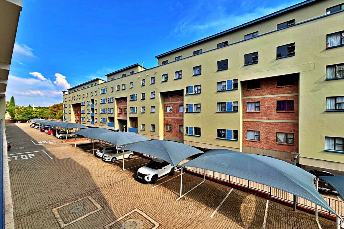 Exterior view of a multi-story apartment complex featuring a paved parking area with shade cloth carports and a brick and plaster facade.