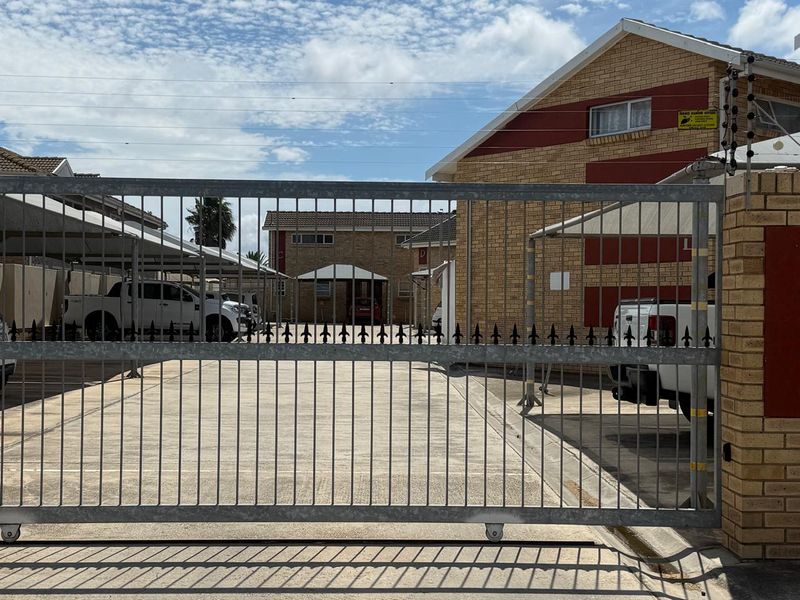 View of the townhouse complex entrance featuring a secure metal gate, paved driveway, and covered carport parking bays.
