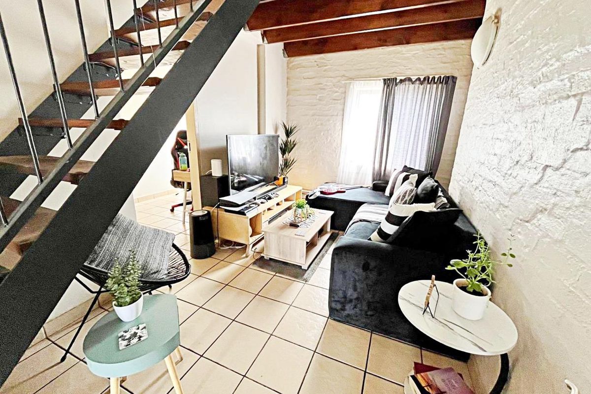 A cozy living room featuring exposed timber ceiling beams, a textured brick wall, and tiled flooring, with an integrated staircase leading to the upper level.
