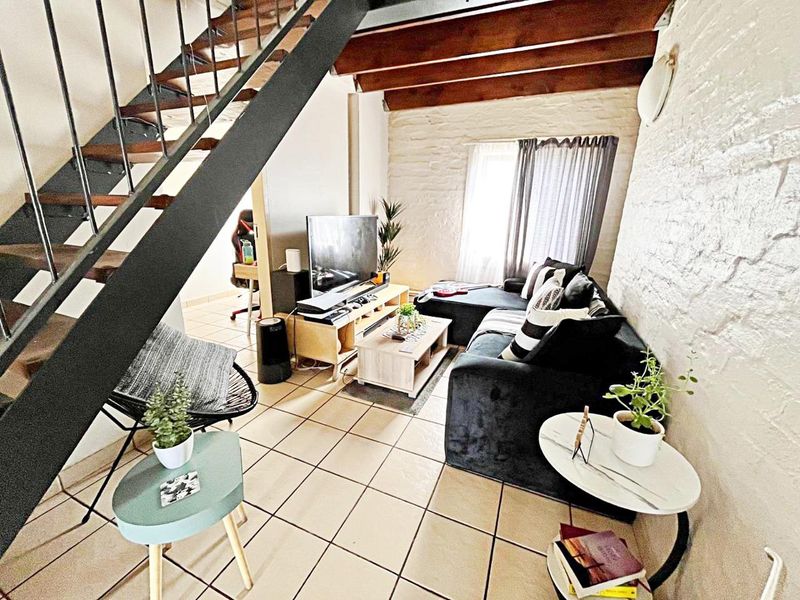A cozy living room featuring exposed timber ceiling beams, a textured brick wall, and tiled flooring, with an integrated staircase leading to the upper level.
