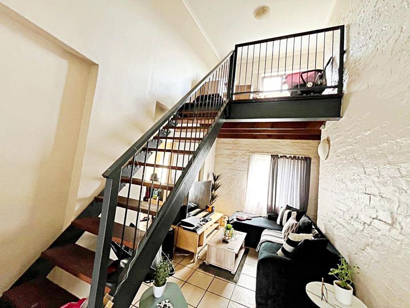 A compact living area featuring a prominent metal staircase leading to a mezzanine level, exposed brick walls, and tiled flooring.