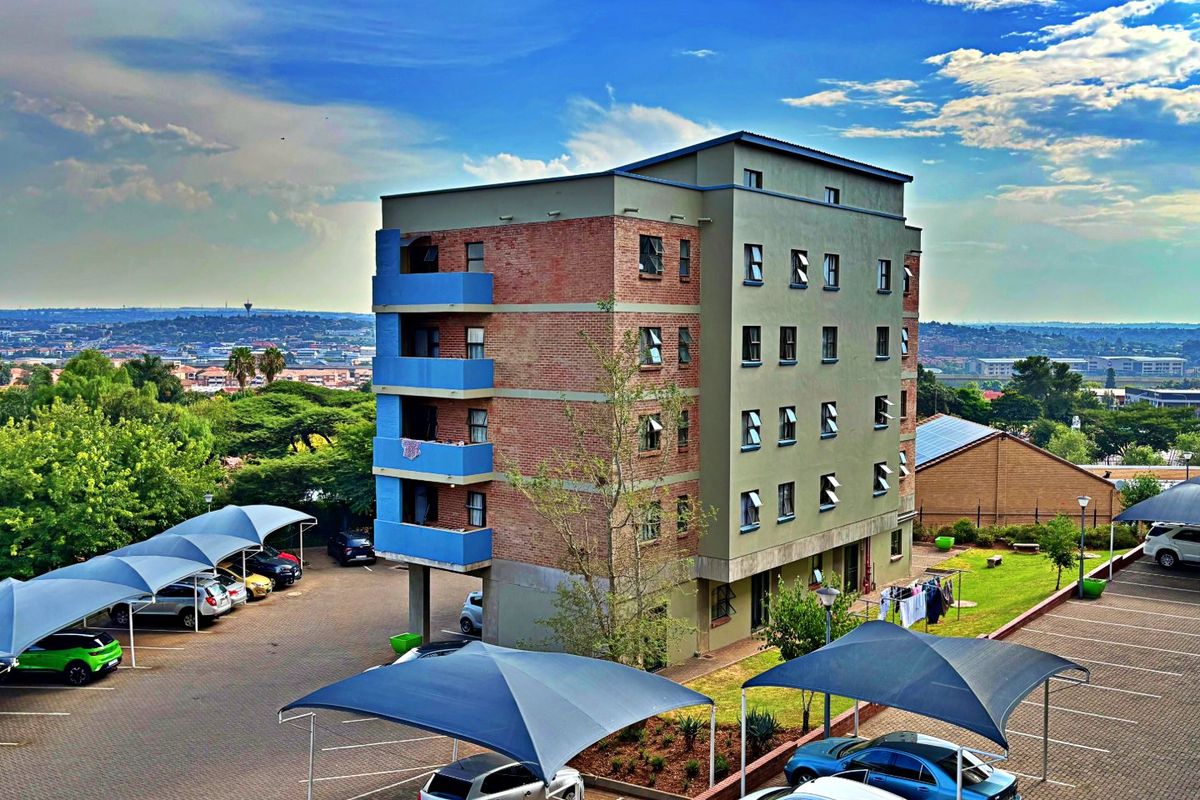 Aerial view of a multi-story apartment building featuring brick and rendered facade, private balconies, and extensive paved parking with shade cloth structures.