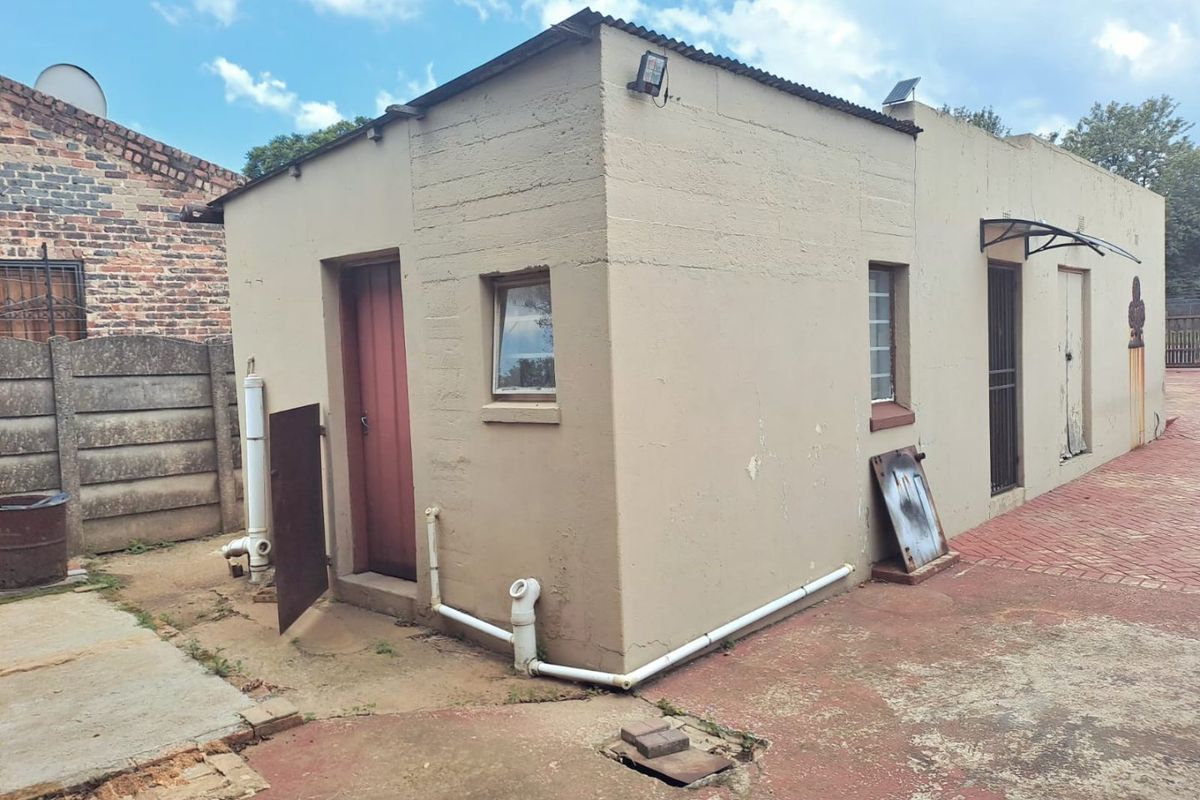 Rear view of a detached outbuilding featuring a corrugated metal roof, security gates, and external plumbing, set on a paved area requiring maintenance.