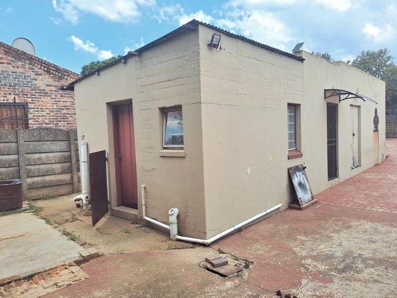 Rear view of a detached outbuilding featuring a corrugated metal roof, security gates, and external plumbing, set on a paved area requiring maintenance.