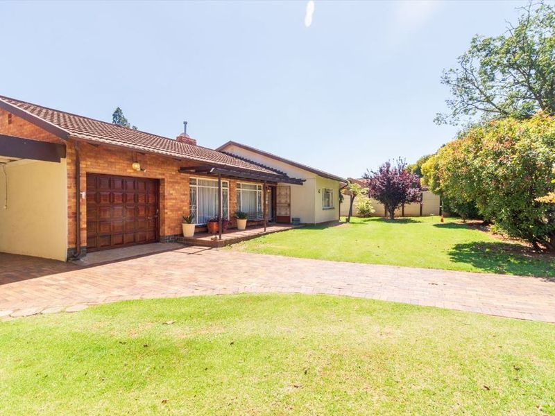 A well-maintained single-story brick house featuring a paved driveway, attached garage, and a spacious front garden with established trees.