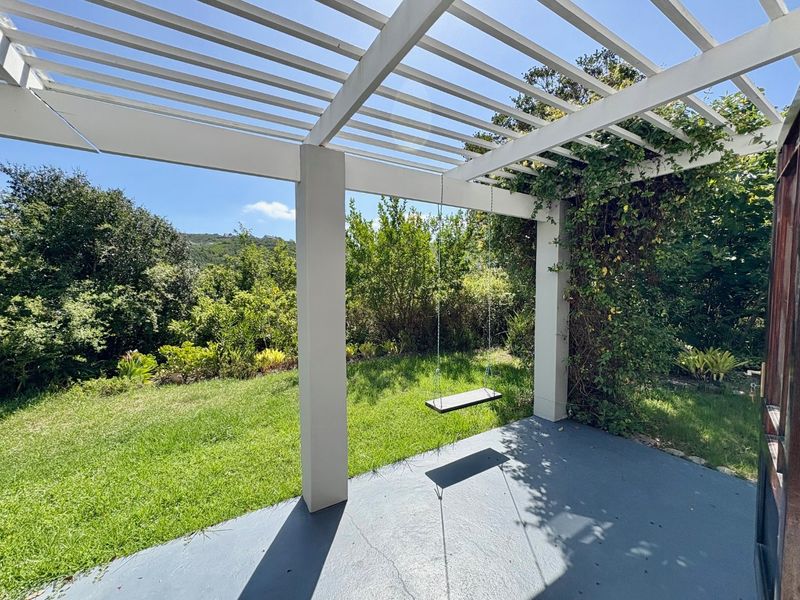 A covered outdoor patio area featuring a white pergola structure and concrete flooring, overlooking a lush green garden.