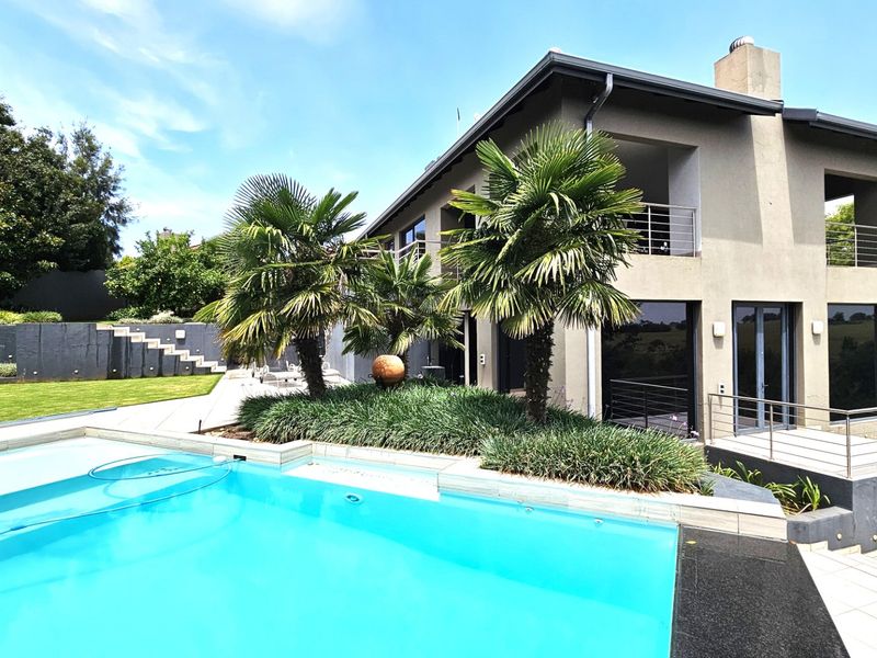Rear view of a modern multi-story home featuring a swimming pool, landscaped garden, tiled patio, and upper-level balcony with stainless steel balustrades.