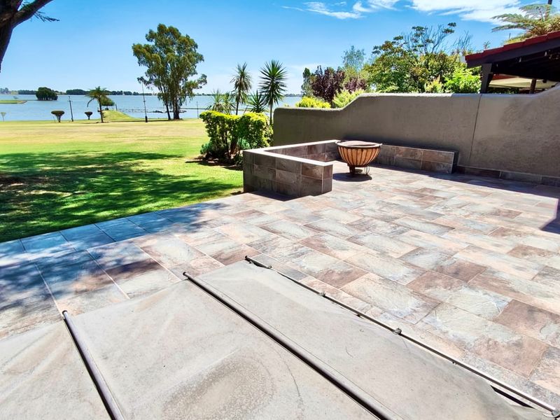 A spacious outdoor patio featuring stone-look tiling, a built-in masonry seating area, and expansive views overlooking a lush green lawn and lake.
