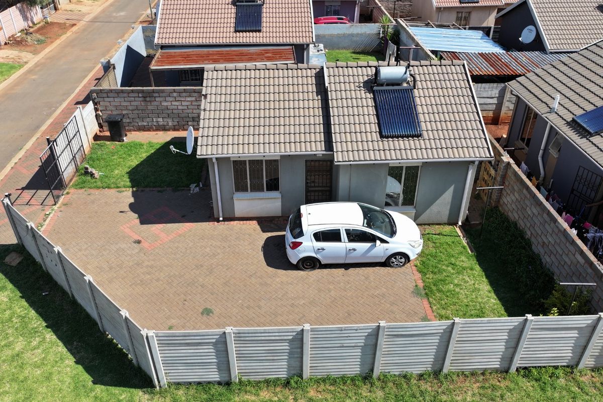 Aerial view of a residential property featuring a tiled roof with a solar water heater, paved driveway, and secure perimeter walling.