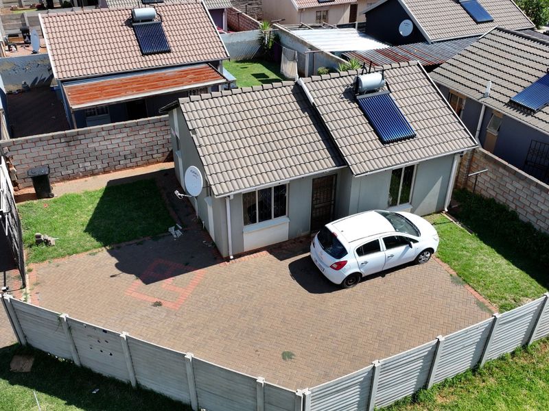 Aerial view of a residential property featuring a tiled roof with a solar water heater, a paved driveway, and a secure boundary wall.