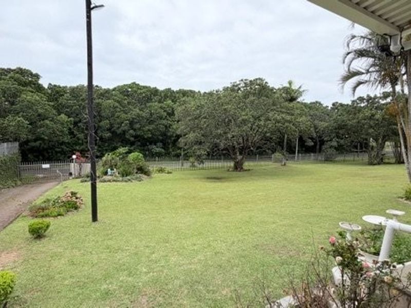 A view from a covered patio overlooking a spacious lawn and garden area with mature trees and perimeter fencing.