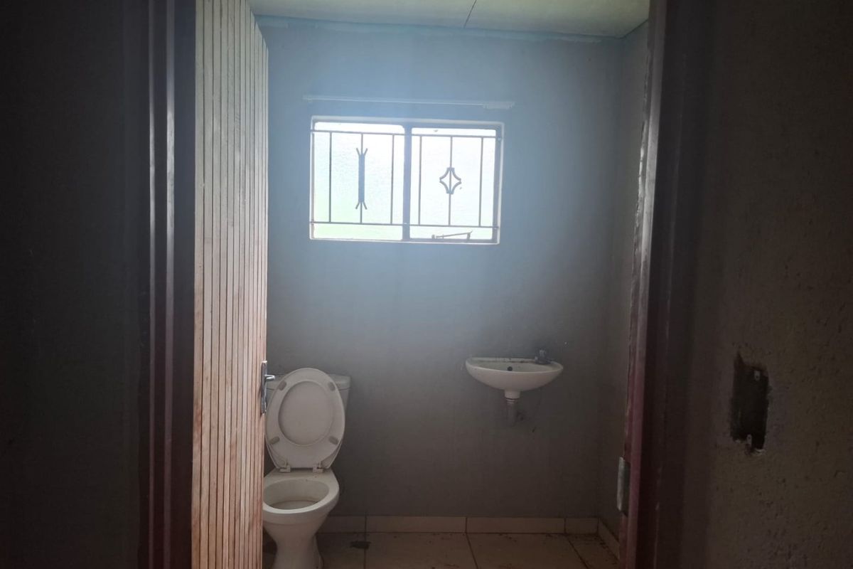 A basic, unfinished bathroom featuring a standard toilet, wall-mounted basin, and security bars on the window, requiring cosmetic updates and finishing touches.