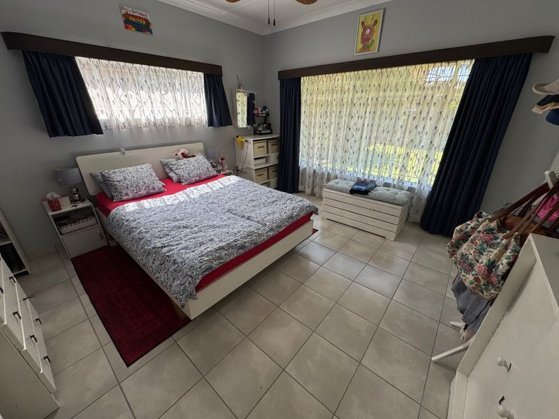 A spacious bedroom featuring large windows with curtains, tiled flooring, and a ceiling fan, offering a bright and functional living space.