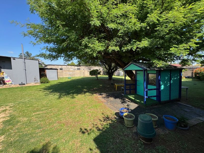 Spacious backyard featuring a large mature tree, a small wooden playhouse, and a paved area, offering a private outdoor space.