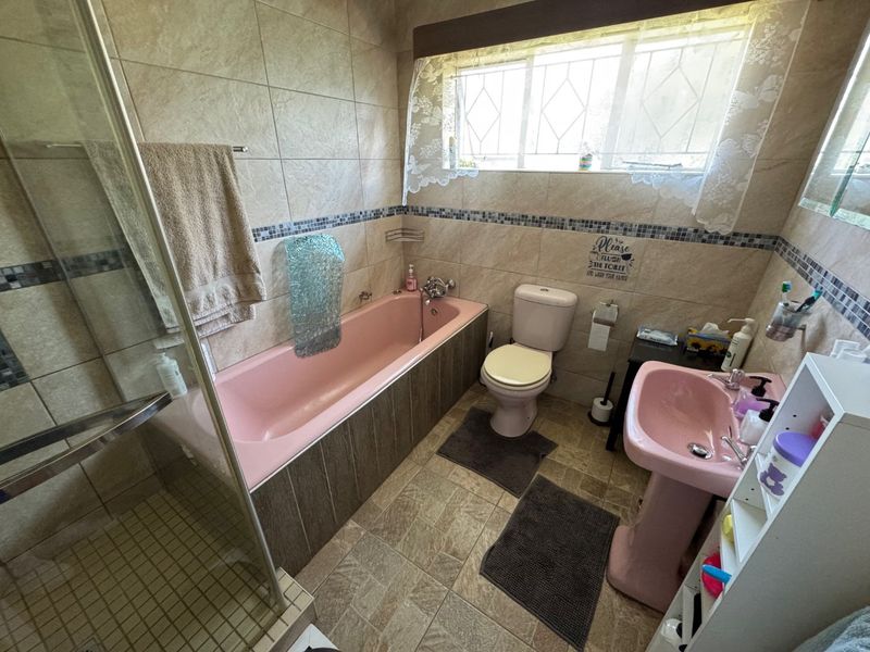A dated bathroom featuring a pink bathtub and pedestal basin, separate glass-enclosed shower, and neutral wall tiling with a decorative mosaic border.