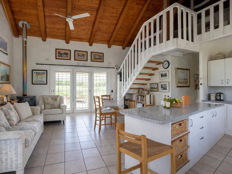 Open-plan living area featuring a vaulted timber ceiling, wood-burning fireplace, tiled flooring, and a mezzanine level accessed by a white staircase.