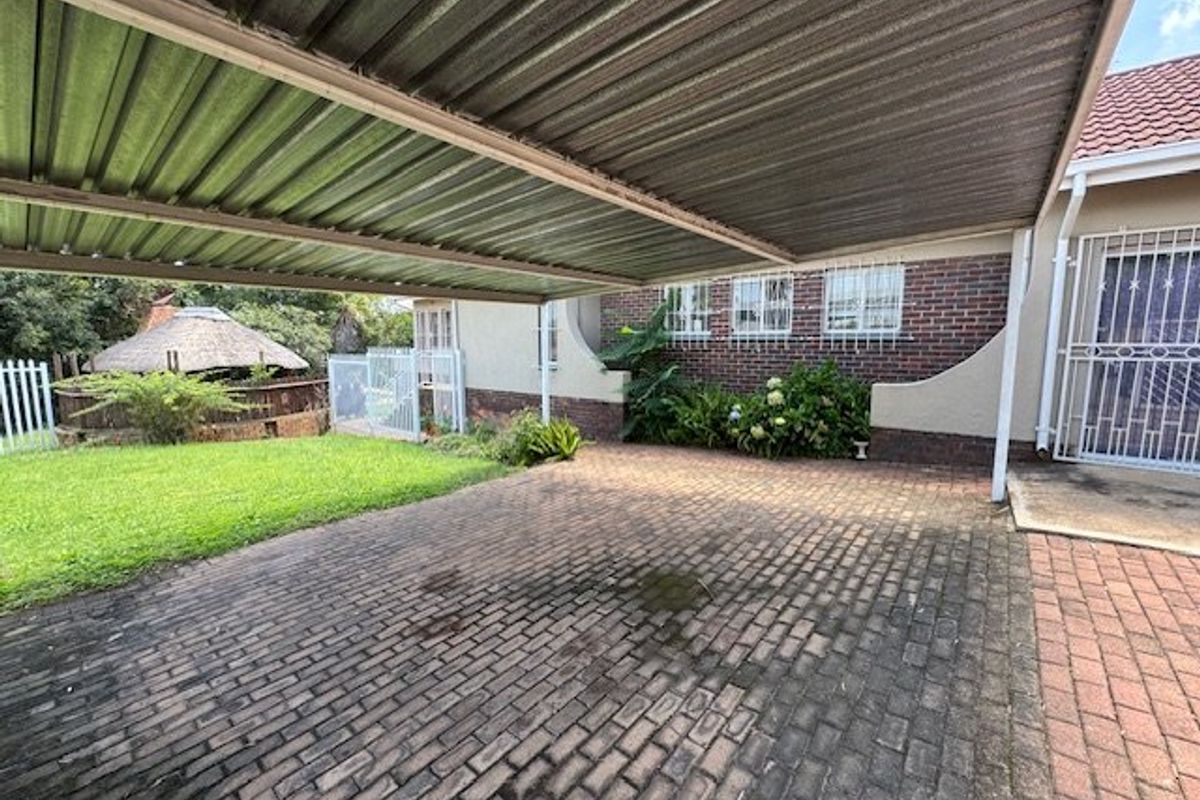 A paved driveway area featuring a large metal carport, brick facade, and security bars on windows, adjacent to a lawn and garden space.