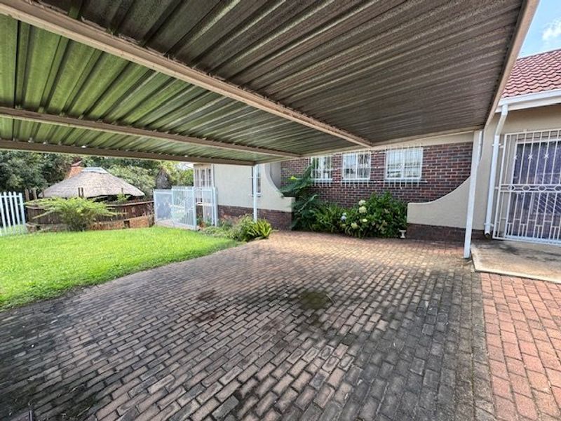 A paved driveway area featuring a large metal carport, brick facade, and security bars on windows, adjacent to a lawn and garden space.