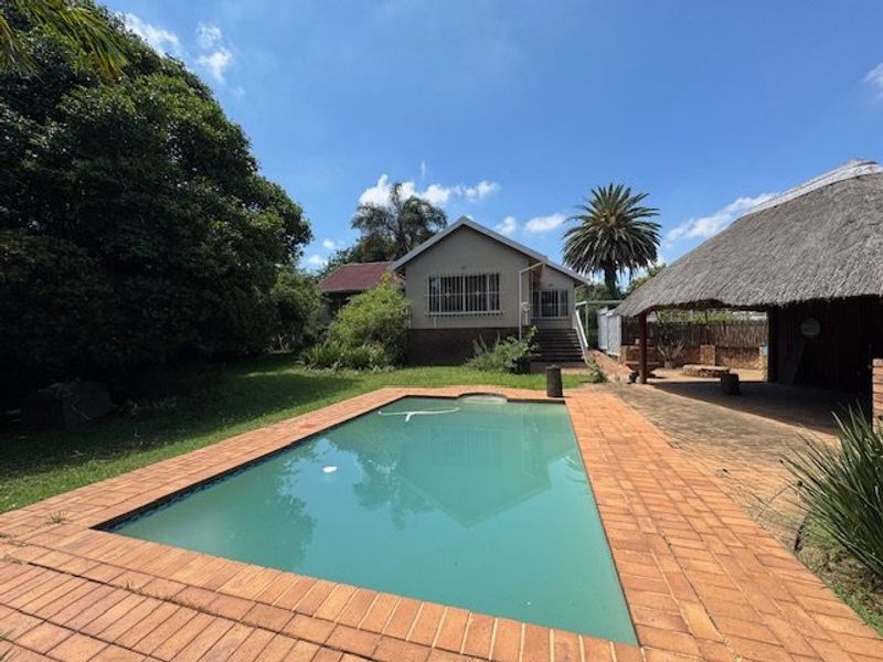 Rear view of the property featuring a swimming pool, paved surround, and a thatched outdoor entertainment area set within a spacious garden.
