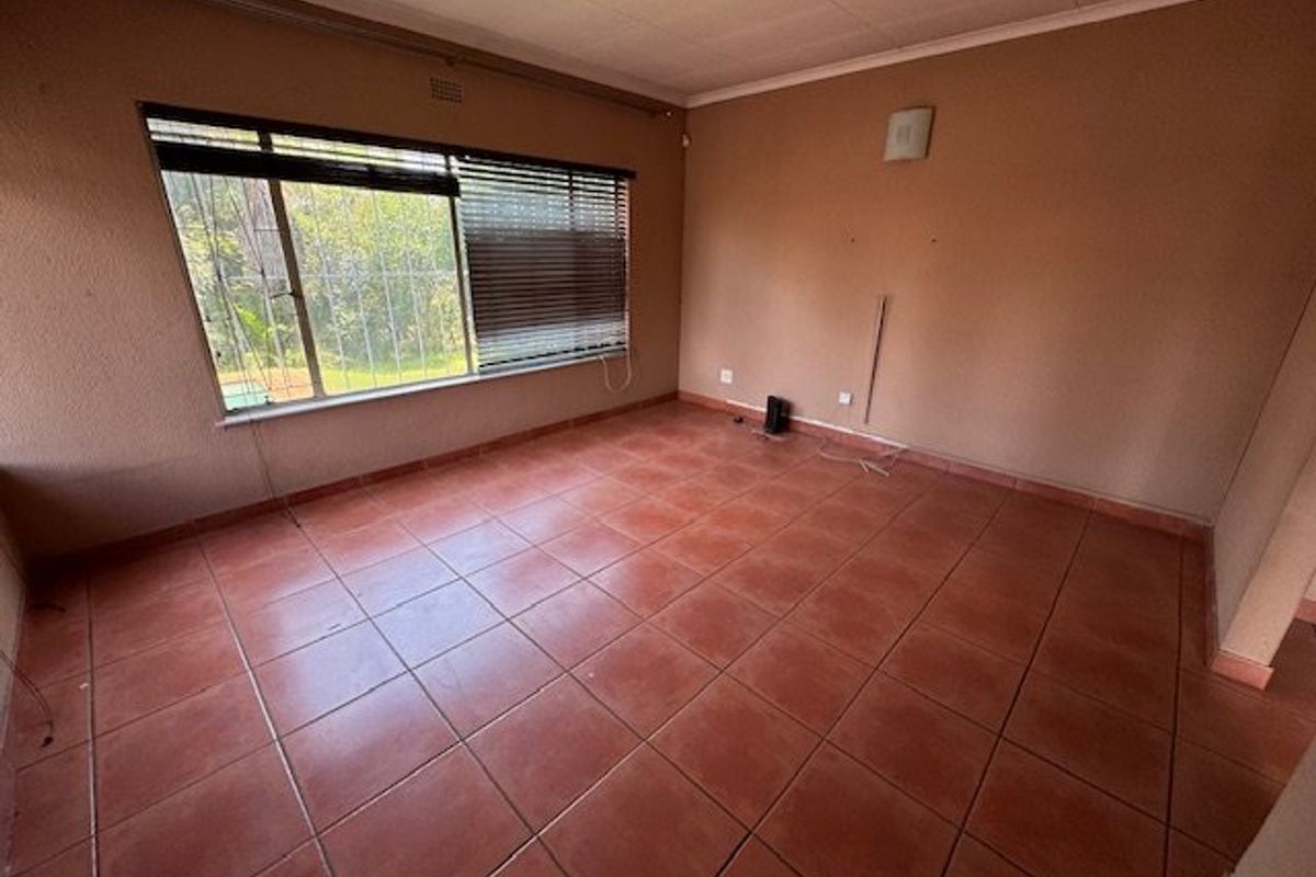 An empty living room featuring terracotta floor tiles, a large window with security bars, and neutral wall finishes.