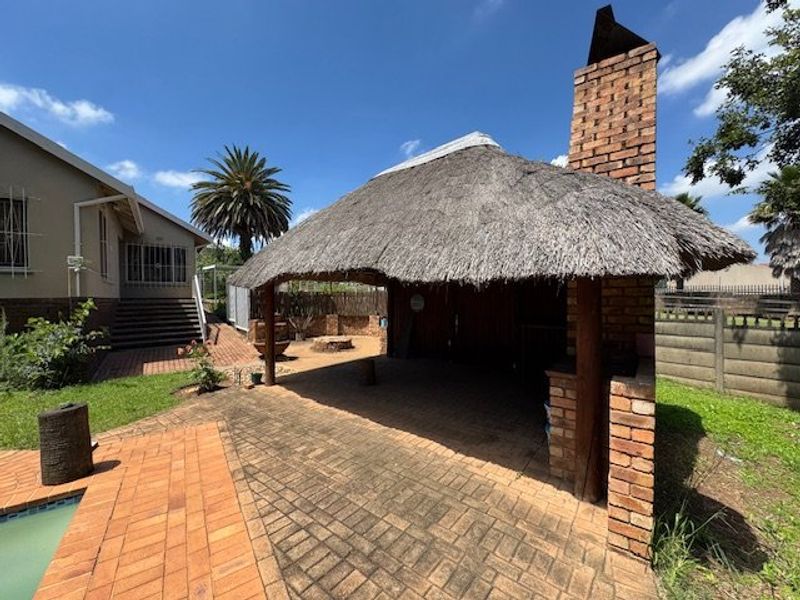 Outdoor entertainment area featuring a traditional thatched lapa with a built-in brick fireplace, adjacent to a paved patio and swimming pool.