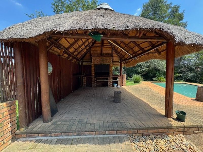 Outdoor entertainment lapa featuring a built-in brick braai, thatched roof, and paved flooring, situated adjacent to the swimming pool area.