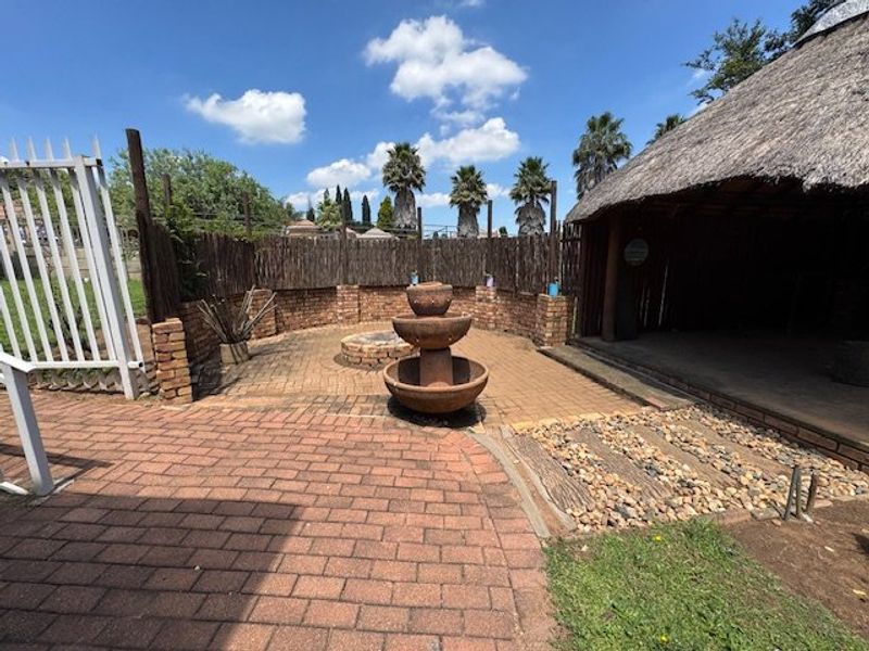 Outdoor entertainment area featuring a paved patio, central water fountain, fire pit, and a traditional thatch-roofed lapa structure.