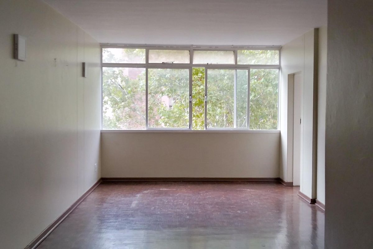 An empty, sunlit living area featuring original parquet flooring and large windows, offering a blank canvas for renovation or interior design.