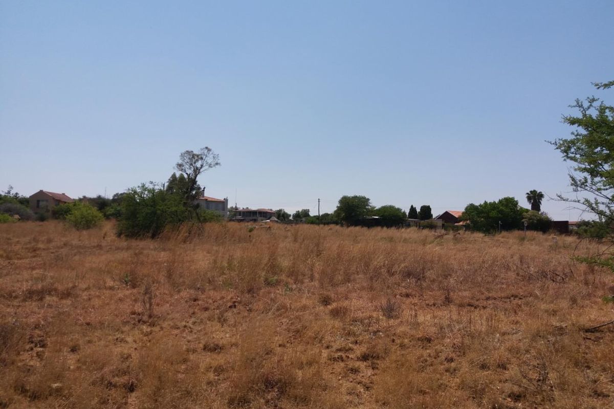 A view of a vacant residential plot featuring dry natural vegetation and surrounding neighborhood properties in the distance.