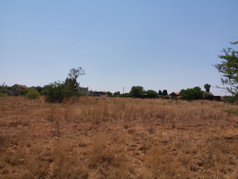 A view of a vacant residential plot featuring dry natural vegetation and surrounding neighborhood properties in the distance.