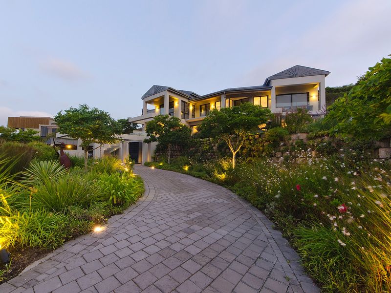 A beautifully landscaped front garden with a paved driveway leading to a modern, multi-level residence featuring exterior lighting and balconies.