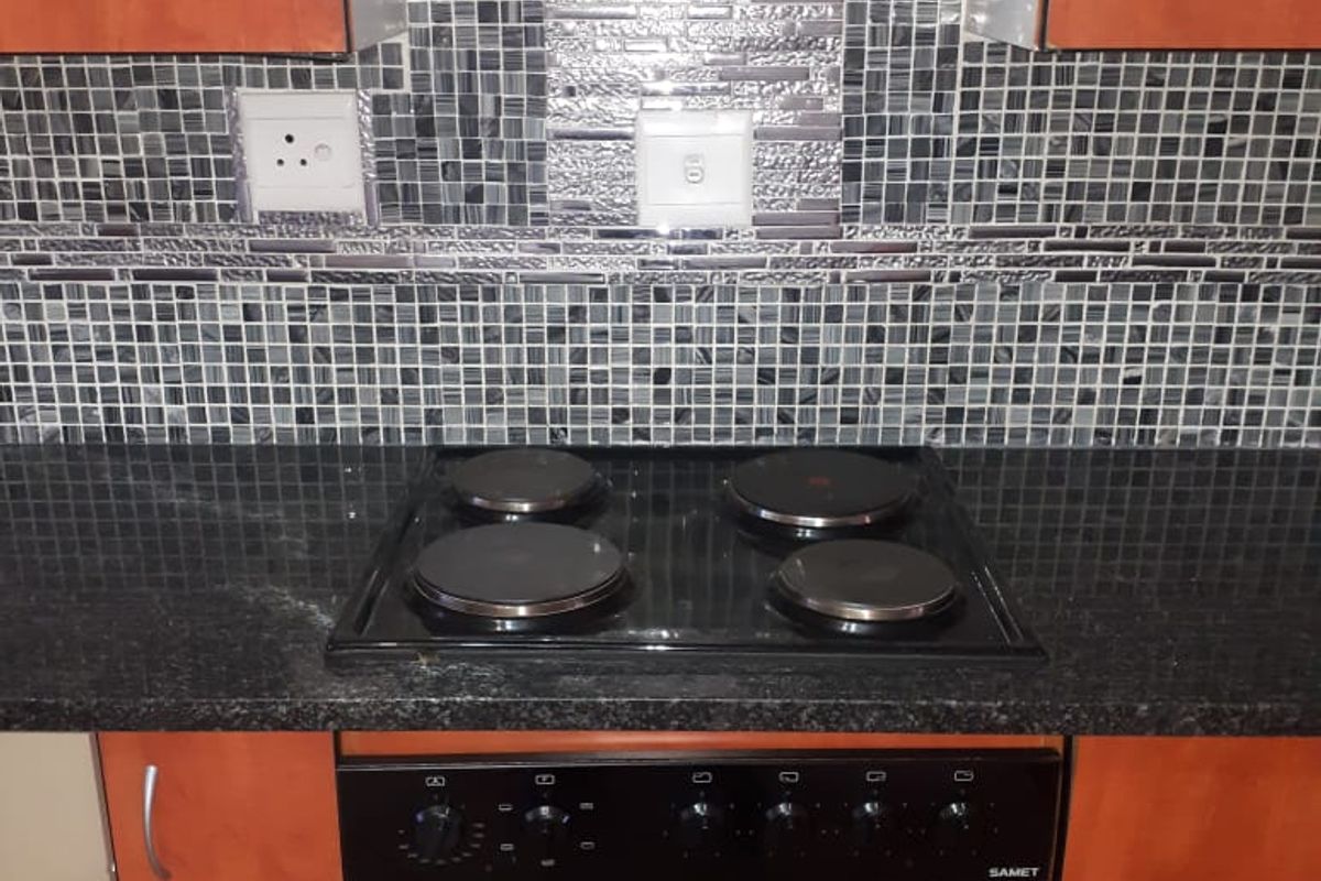 A kitchen workspace featuring a built-in electric hob, oven, extractor fan, and decorative mosaic tile splashback with granite-look countertops.