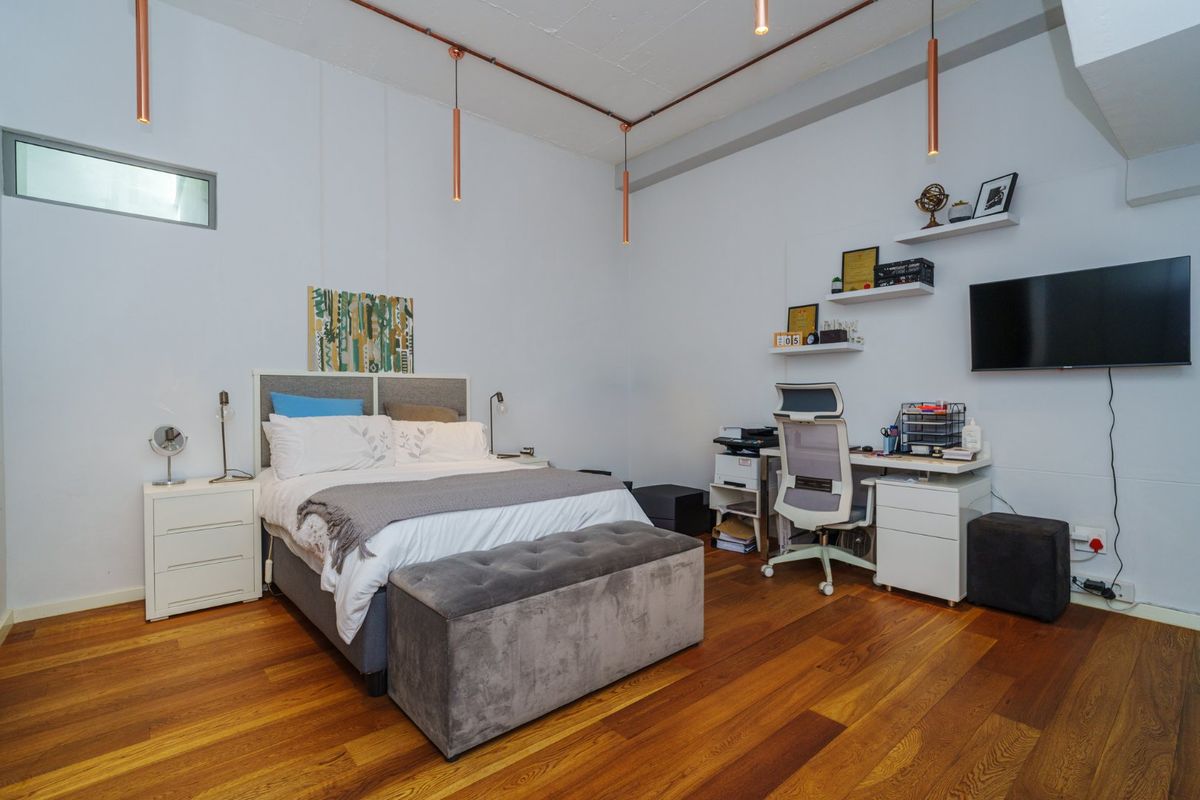 A spacious bedroom featuring polished timber flooring, exposed industrial-style ceiling conduit, and modern pendant lighting.