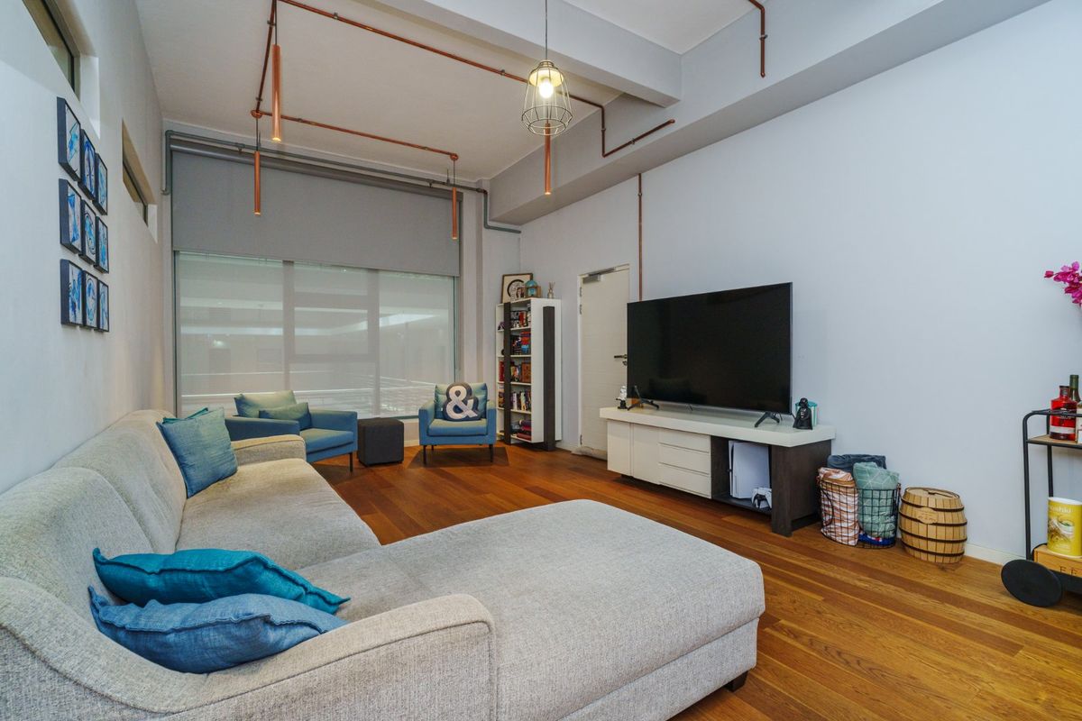 A modern living room featuring exposed industrial-style copper piping, hardwood flooring, and large windows with roller blinds.