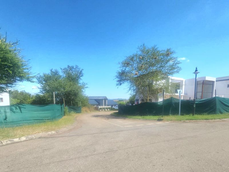 A street-level view of a residential development site featuring paved roads, mature trees, and temporary construction fencing surrounding the property.