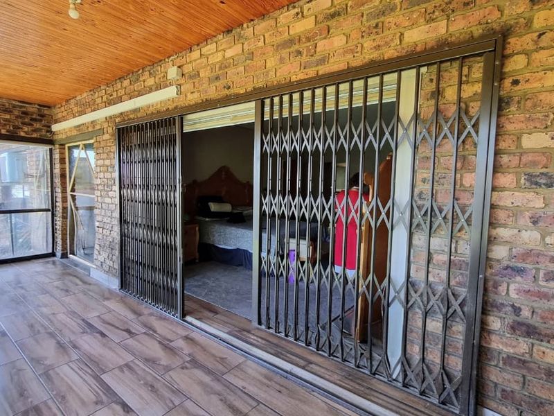 A covered patio area featuring wood-look tiled flooring, exposed brick walls, and a timber-clad ceiling, providing access to the interior through security gates.