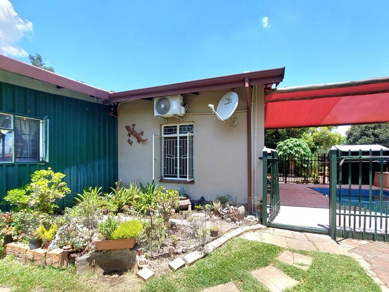 Rear exterior view featuring a paved patio area, swimming pool, shade sail, and established garden beds with security features.