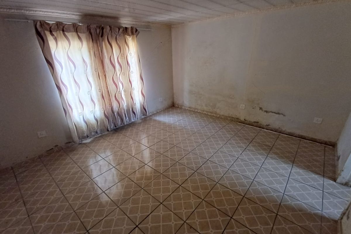 An empty bedroom featuring tiled flooring and a window with curtains, showing signs of significant wall damp and surface deterioration requiring maintenance.