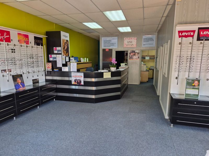 Commercial retail space featuring a central reception desk, built-in eyewear display shelving, carpeted flooring, and a suspended ceiling with integrated lighting.