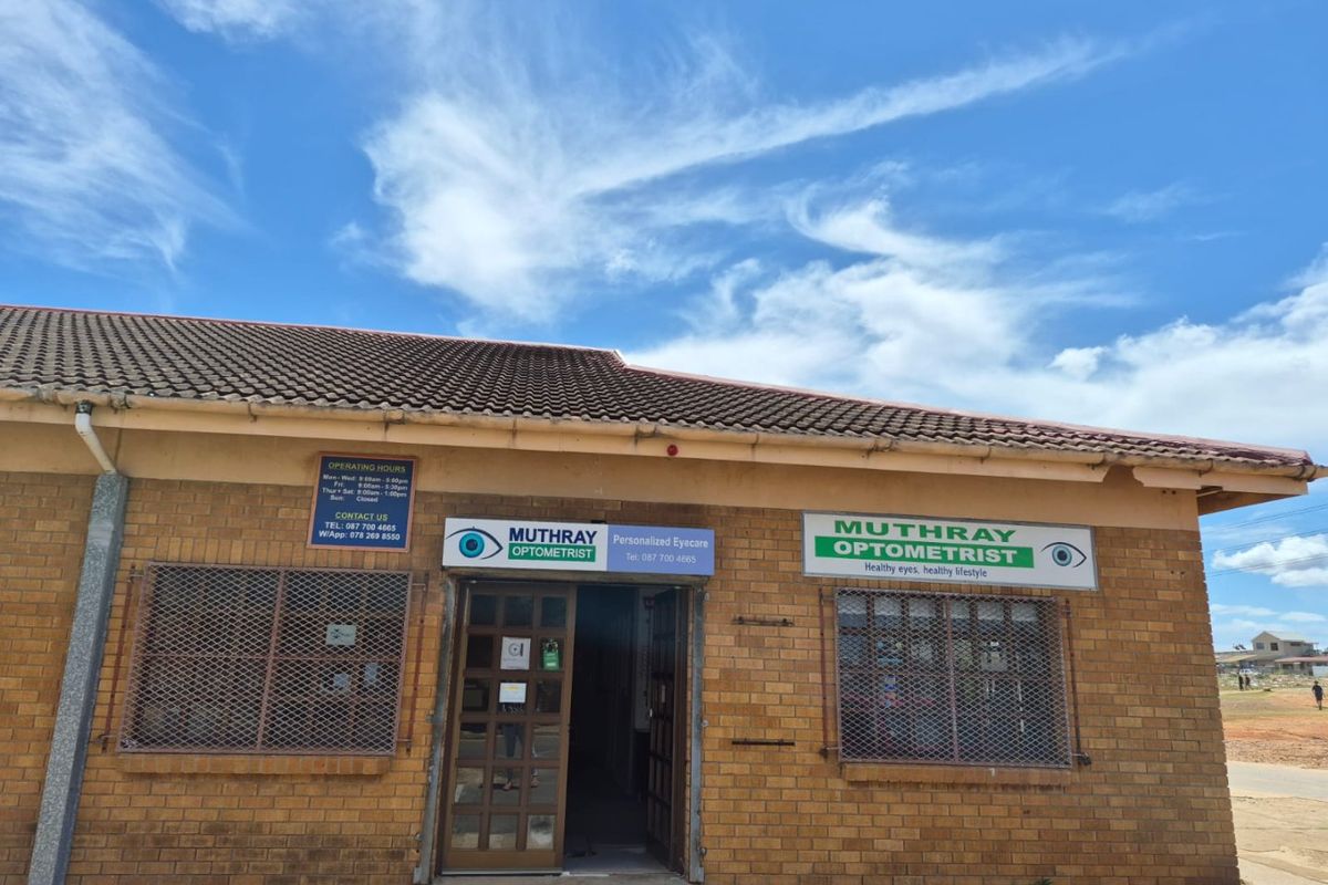 Front facade of a commercial optometrist practice featuring brick construction, tiled roofing, and prominent business signage with security grilles on windows.