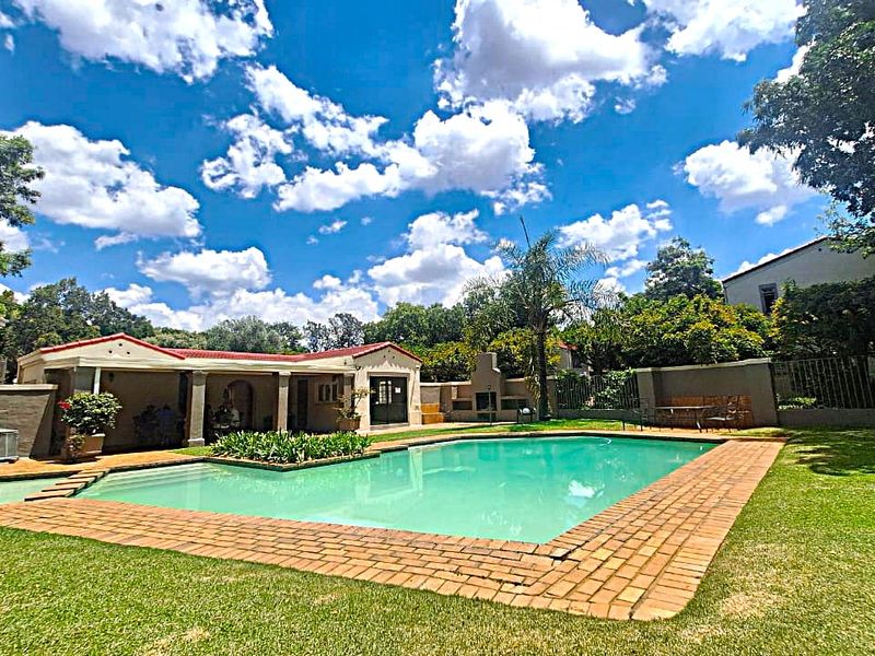 A spacious outdoor pool area featuring a swimming pool with brick coping, a covered patio, and a built-in braai set within a landscaped garden.