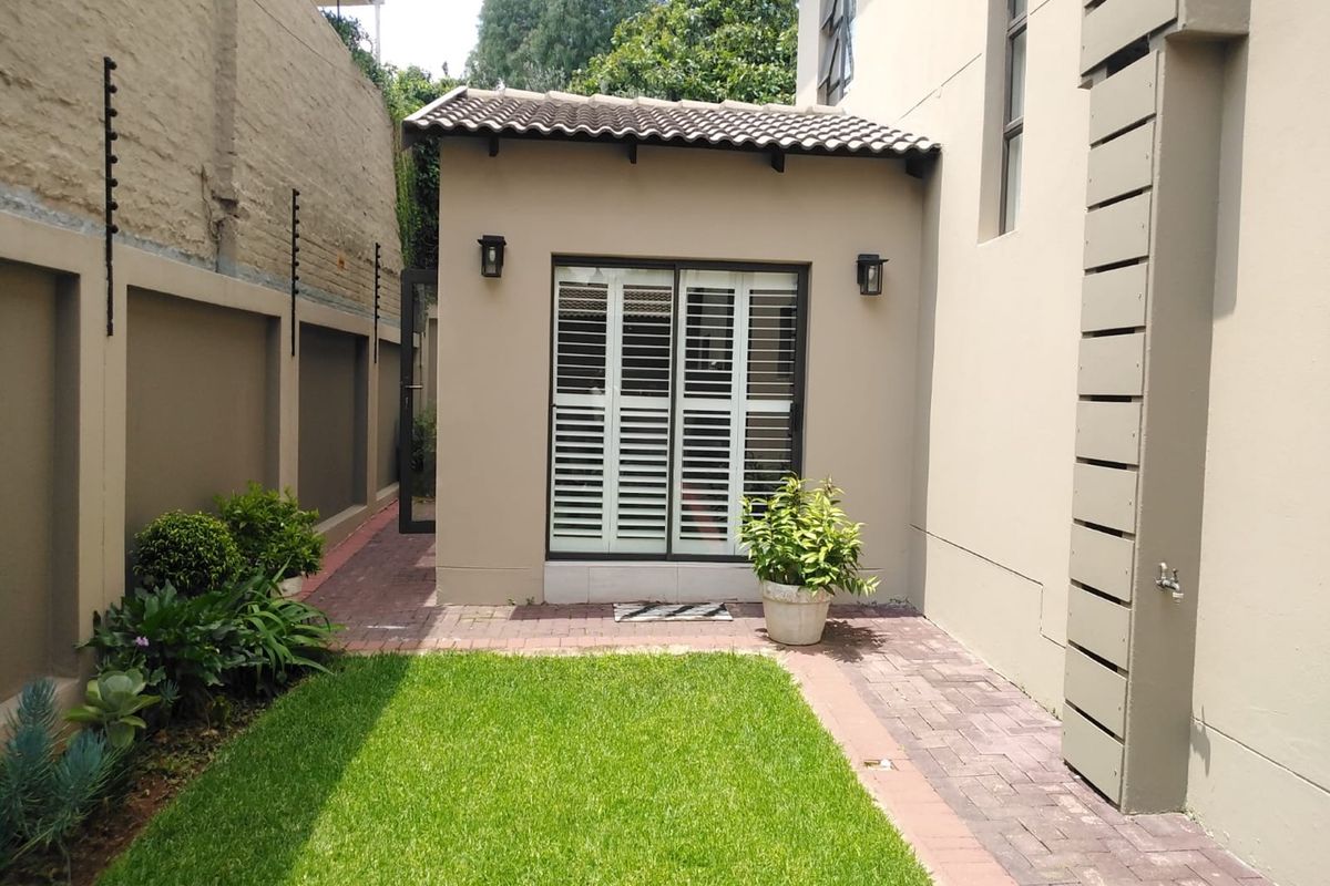 A well-maintained rear exterior area featuring a paved walkway, manicured lawn, boundary wall with electric fencing, and a sliding door with security shutters.